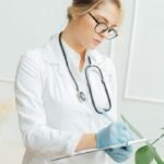Female doctor with eyeglasses taking notes in a hospital, wearing a stethoscope and gloves.
