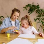 A mother and daughter engage in creative finger painting at a table indoors.