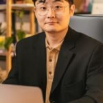 Confident Asian businessman in eyeglasses working on a laptop with startup note, indoors.